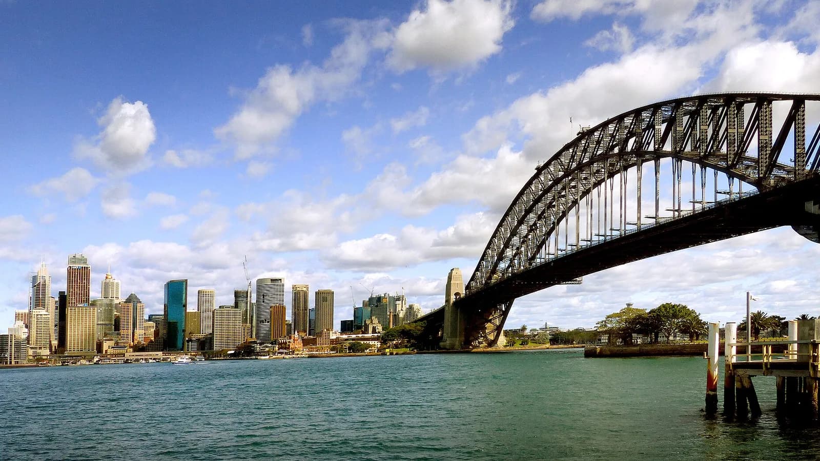 The Sydney central business district skyline across the harbour, with the steel arch of the Sydney Harbour Bridge on the right under a partly cloudy sky.