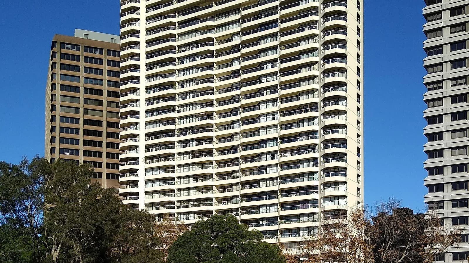 Tall pale residential apartment tower of The Connaught in central Sydney, with balconies rising above street-level shops and smaller brick buildings under a bright blue sky.