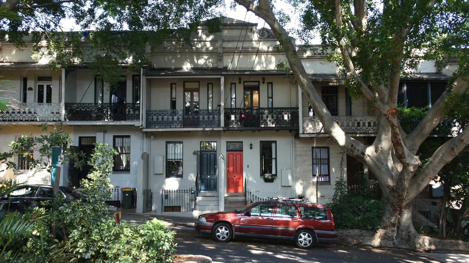 A row of two-storey terrace houses with cast-iron balcony railings and a red front door, shaded by a large tree, with a red station wagon parked in the street in front.