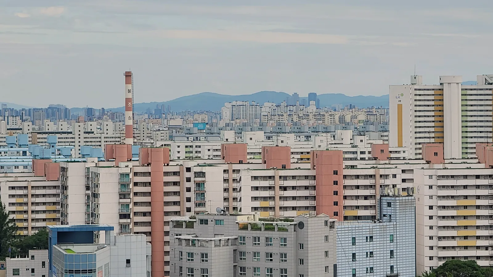 Rows of white and cream high-rise apartment blocks with salmon-pink stair towers fill the view, with a red-and-white chimney, more housing and low hills on the horizon under a grey sky in Nowon-gu, Seoul.