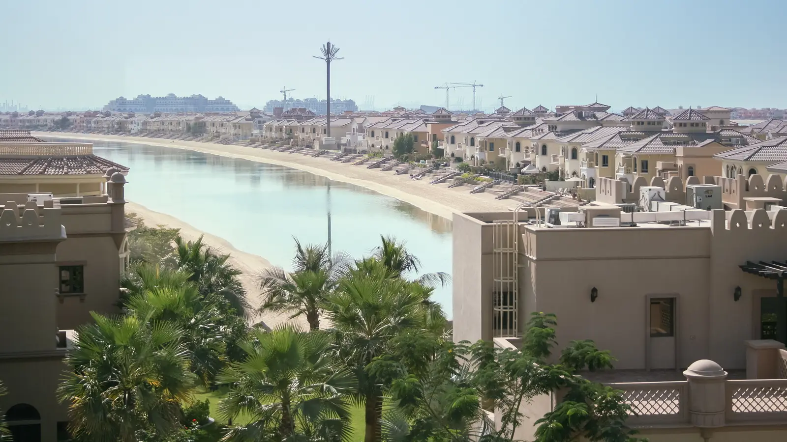 Rows of beige villas along a sandy lagoon shore on a frond of Palm Jumeirah, Dubai, with palm trees in the foreground.
