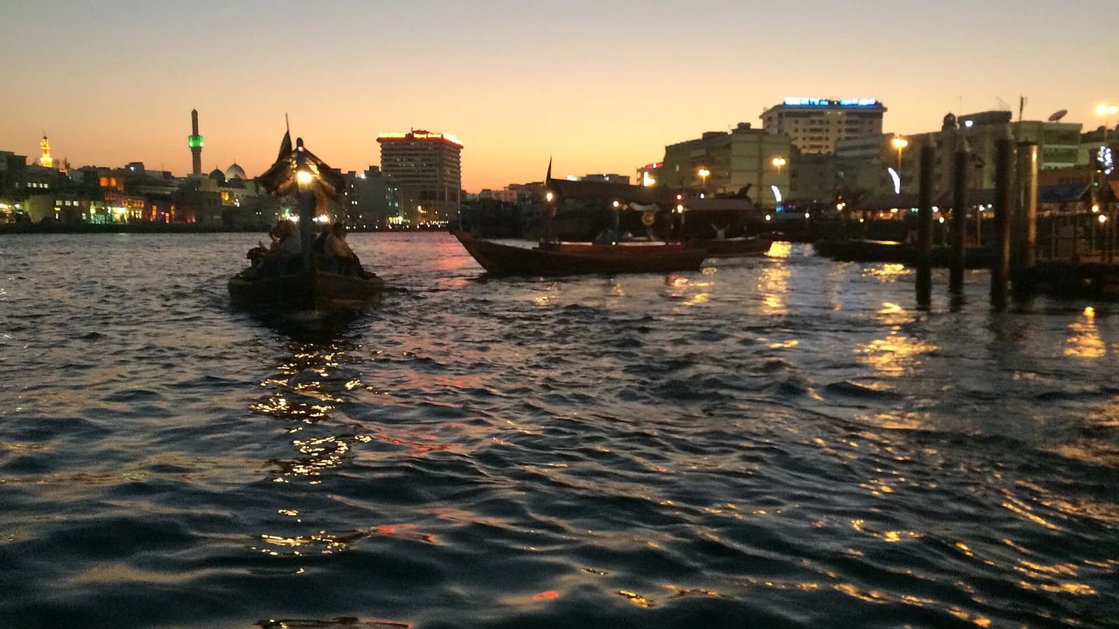 Wooden abras cross Dubai Creek at dusk, with the Deira waterfront skyline, minarets and lit buildings reflecting on the water.