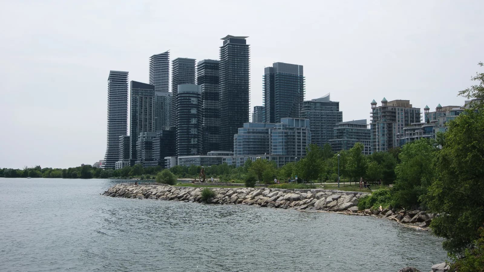 Glass condominium towers at Humber Bay on Toronto's western waterfront, seen across Lake Ontario from a rocky shoreline with trees in the foreground under a pale sky.