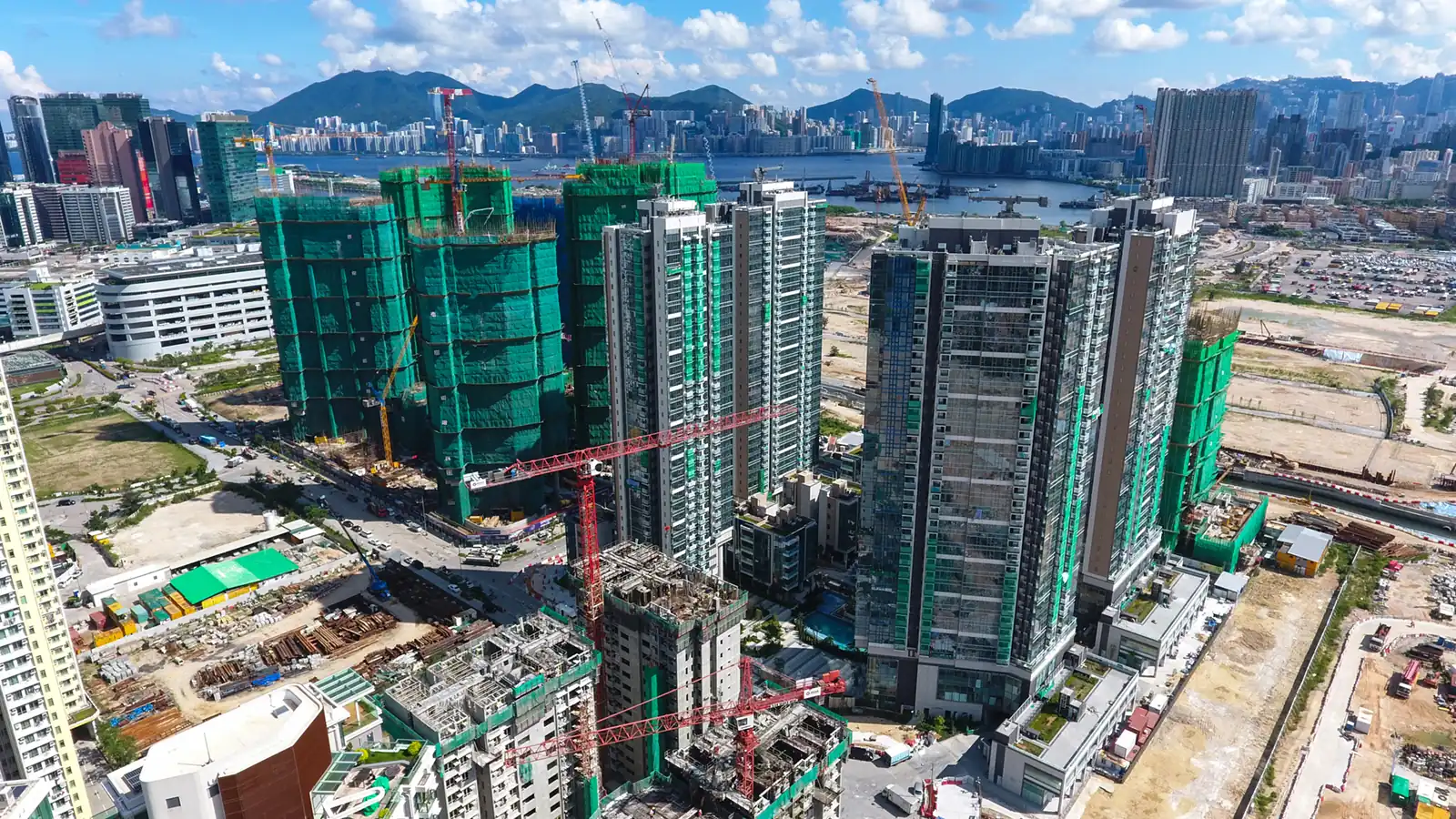 Residential towers under construction, several wrapped in green netting, with tower cranes on the former Kai Tak airport site in Kowloon, Hong Kong, and Victoria Harbour and hills behind.