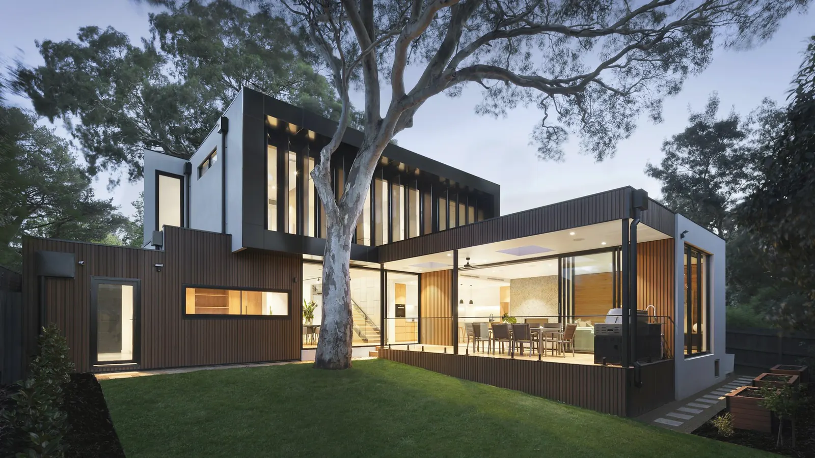 A contemporary house at dusk, with warm light in the rooms and a long table under a timber roof.