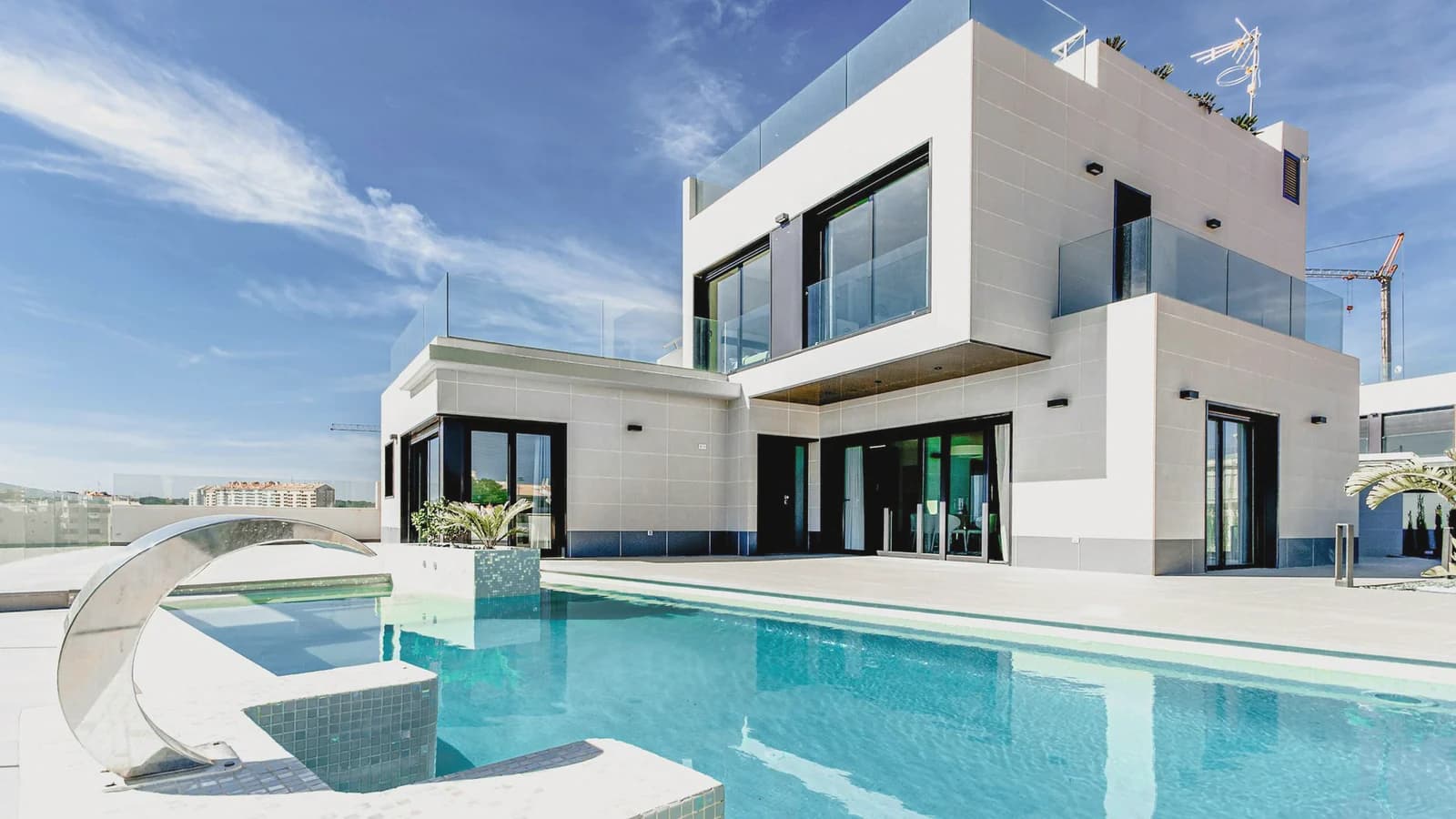 A white modern house in daylight, with a turquoise pool and lounge chairs along the water.