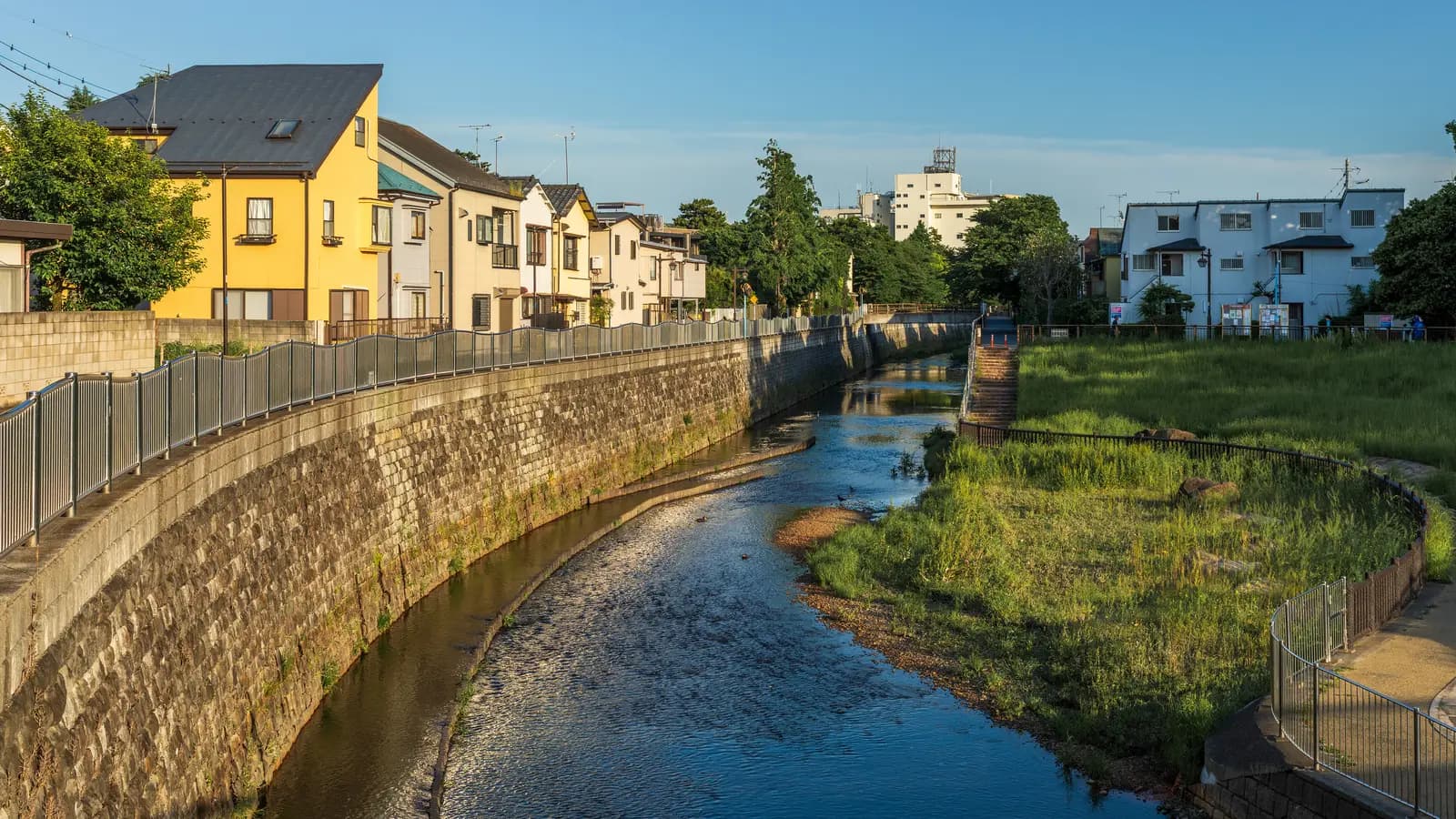 Two- and three-storey houses and a low apartment block line a stone-walled stretch of the Zenpukuji River in a Tokyo residential neighbourhood under a clear evening sky.
