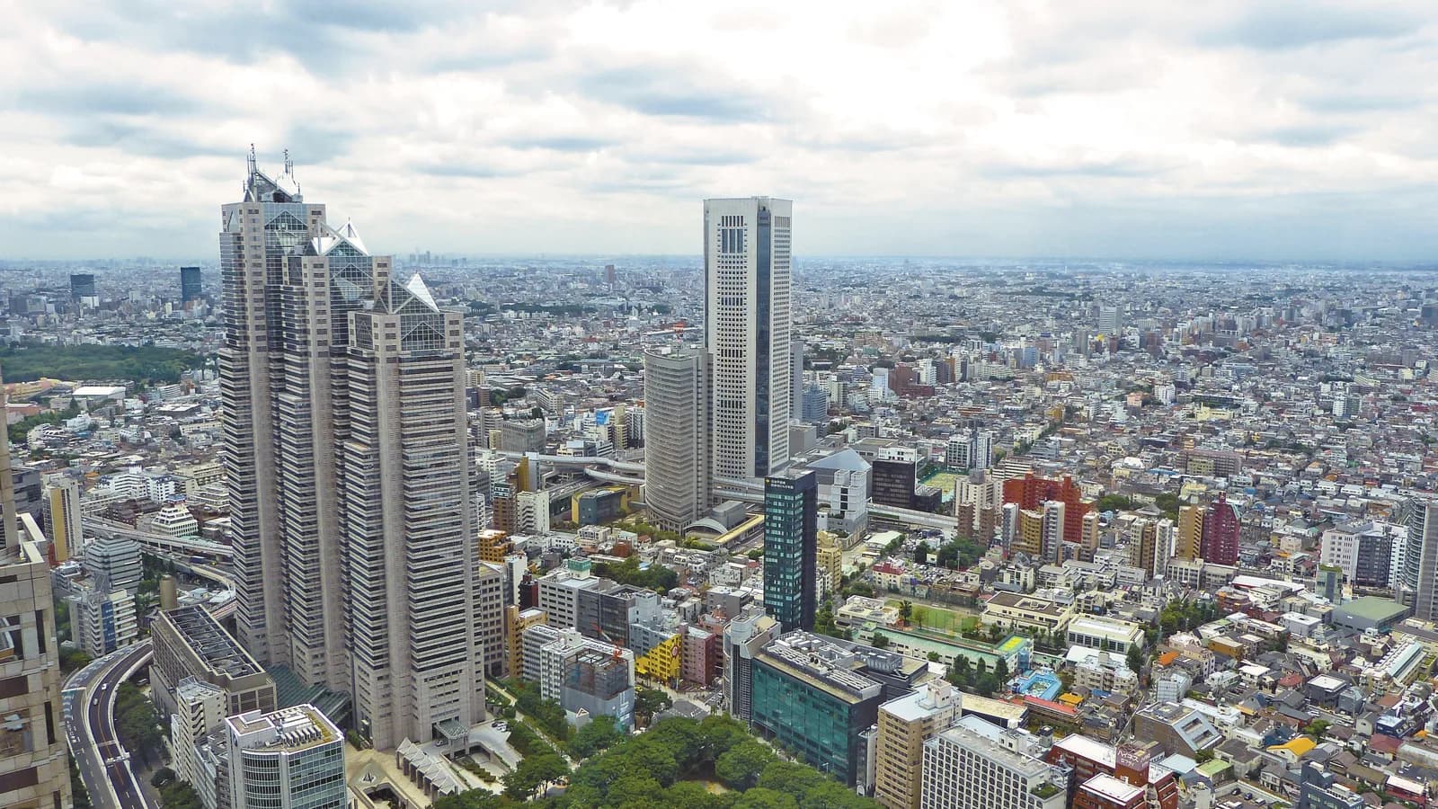 High-angle view over central Tokyo on an overcast day, with the Shinjuku Park Tower skyscrapers on the left, a slim tall tower in the centre, and dense low-rise districts stretching to the horizon.