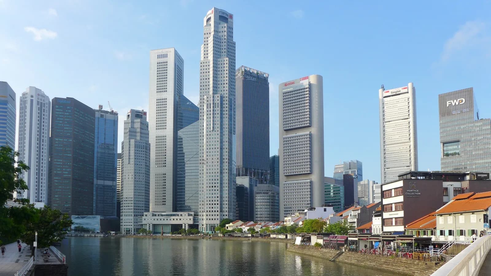 Office towers of Singapore's Raffles Place financial district rising above the Singapore River and low-rise shophouses at Boat Quay, under a clear sky.