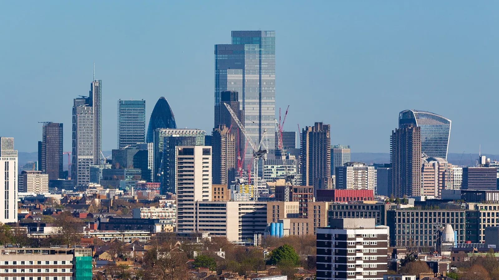 The office towers of the City of London rise behind rooftops, terraced houses and blocks of flats in north London under a clear blue sky, seen from Parliament Hill.