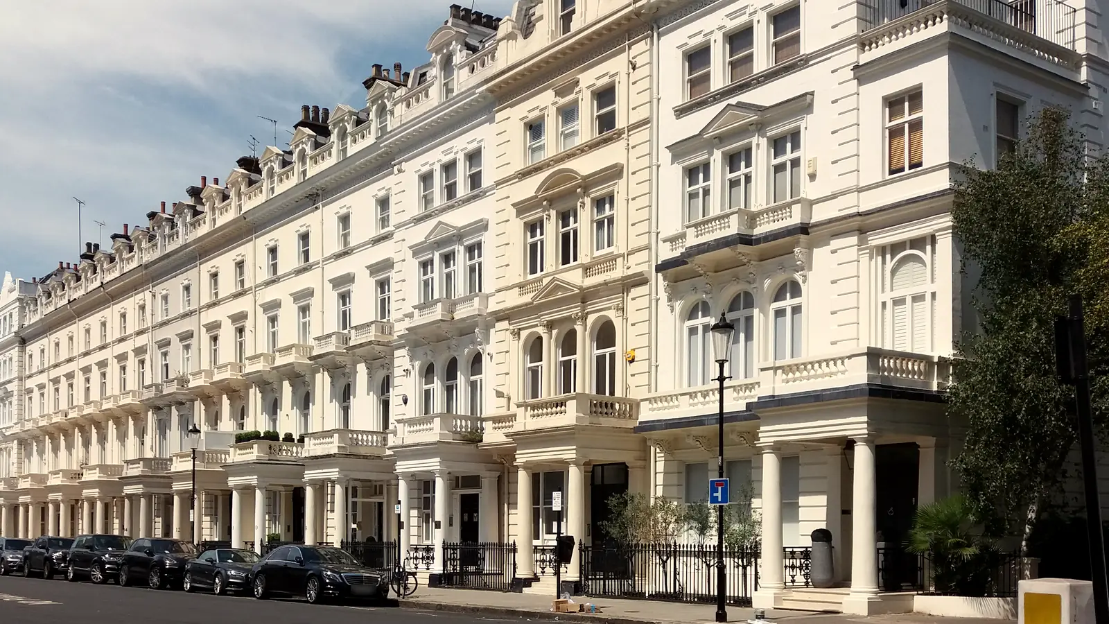 A long row of white stucco terraced houses on Queen's Gate Terrace, South Kensington, with columned porches, first-floor balconies and cars parked along the kerb under a blue sky.