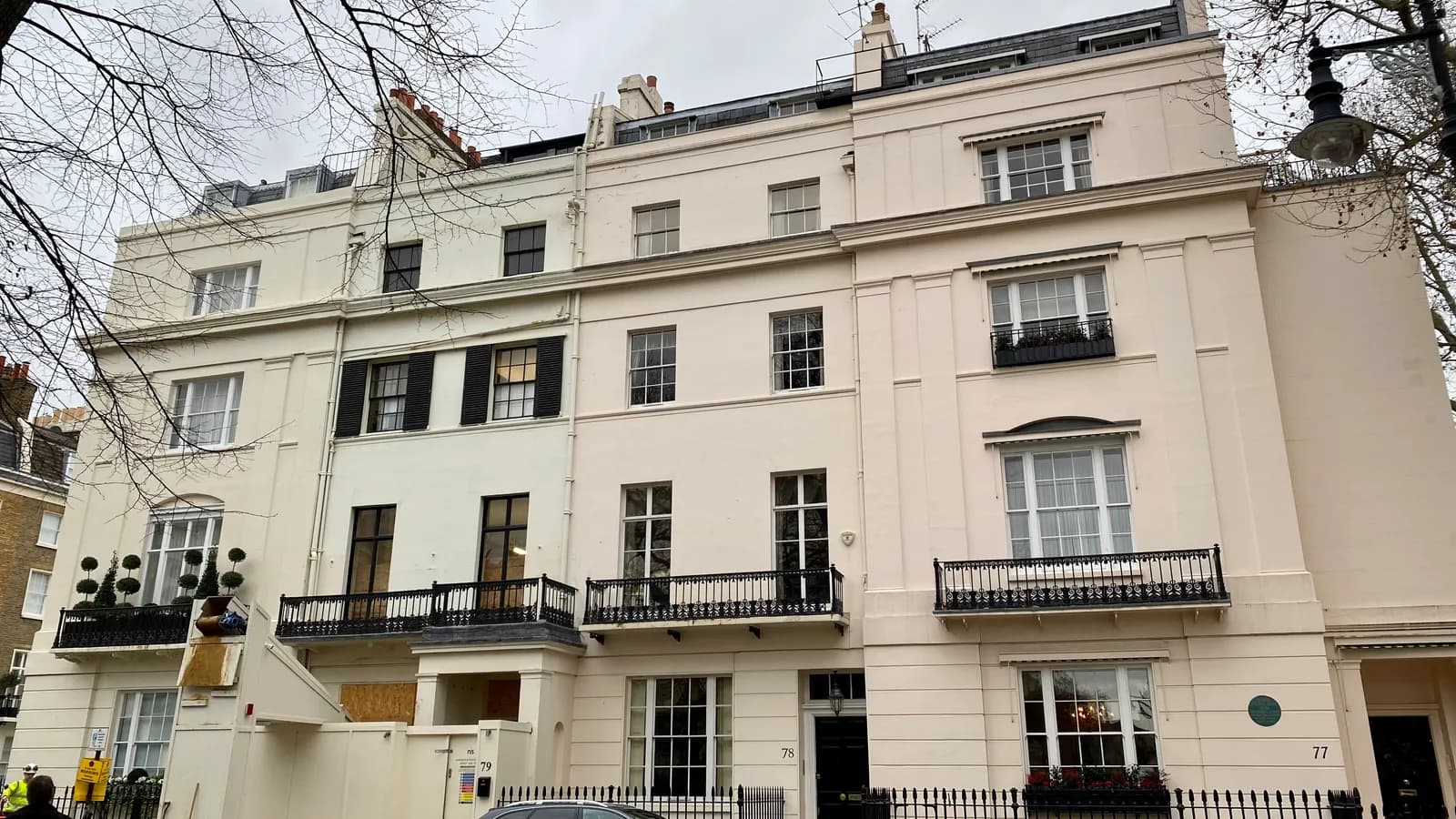 A terrace of white stucco houses on Chester Square in Belgravia, London, four and five storeys high, with black iron balconies on the first floor, black railings along the pavement and a car parked in front, under a grey winter sky.