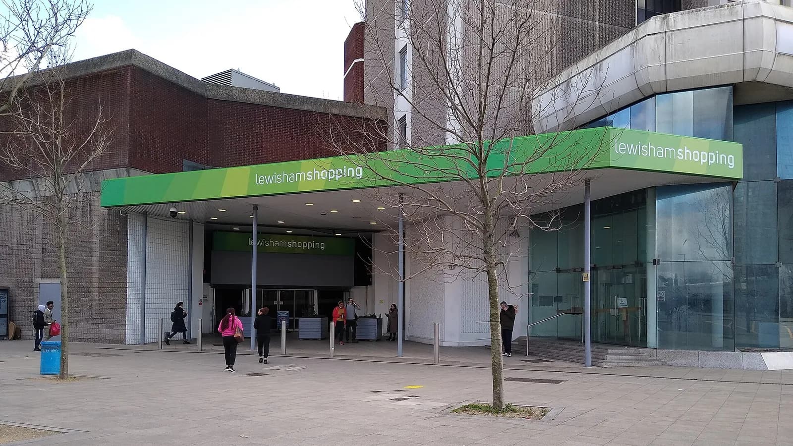 Entrance to Lewisham Shopping Centre in south-east London, with a green canopy reading lewishamshopping over the doors, a brick and concrete 1970s block behind, bare trees and shoppers walking across a paved square.