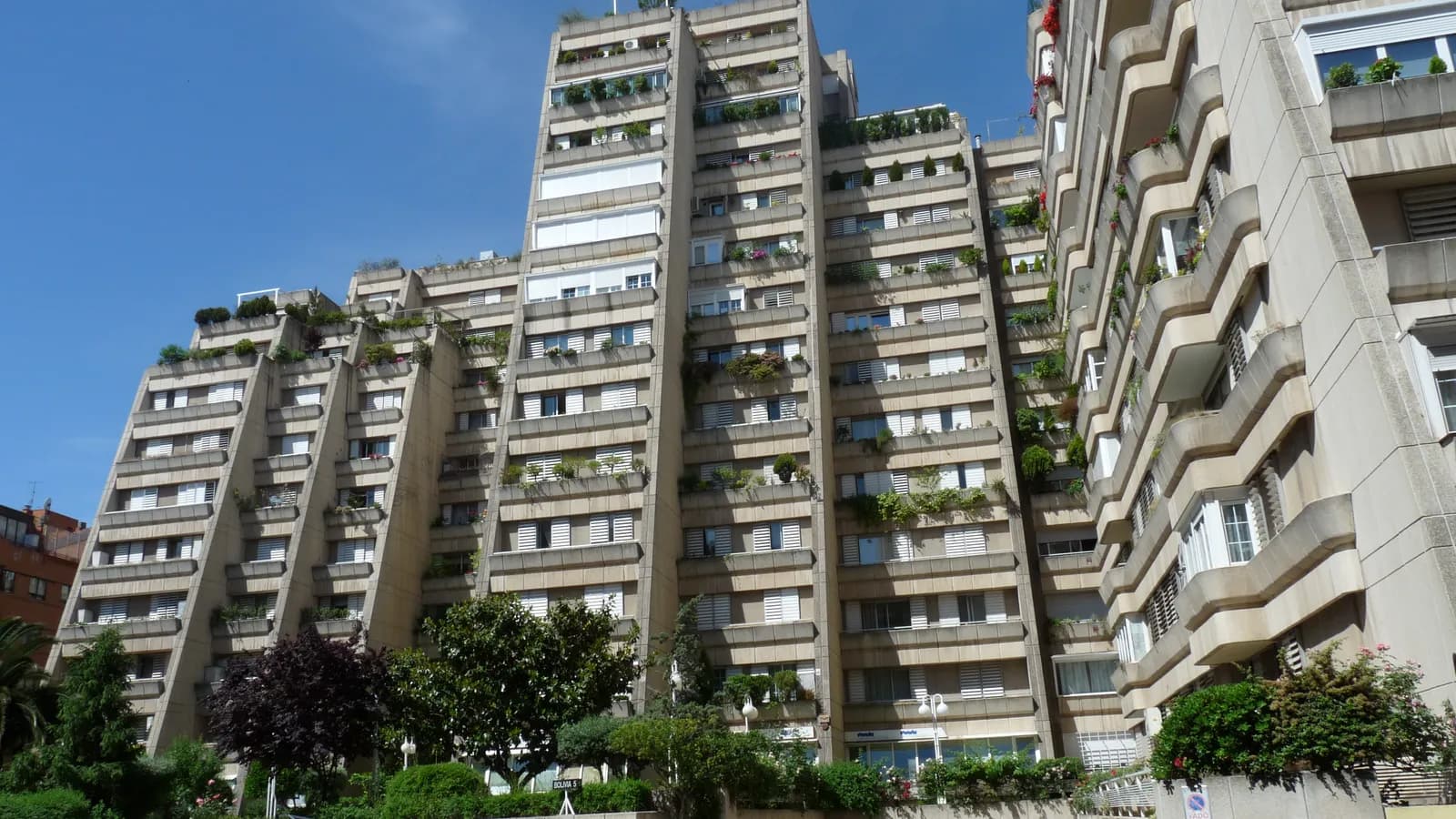 Tall stepped blocks of flats in beige concrete on Calle Bolivia in Madrid, with rows of balconies full of plants and shutters, behind trees and a low wall, under a clear blue sky.