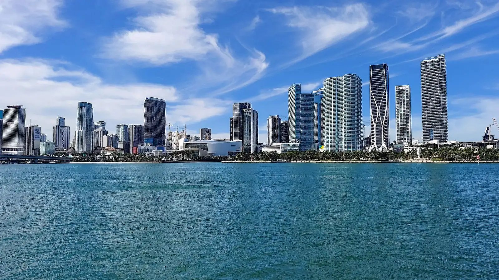 The towers of Downtown Miami and the Biscayne Bay waterfront seen across turquoise water under a blue sky with thin clouds.