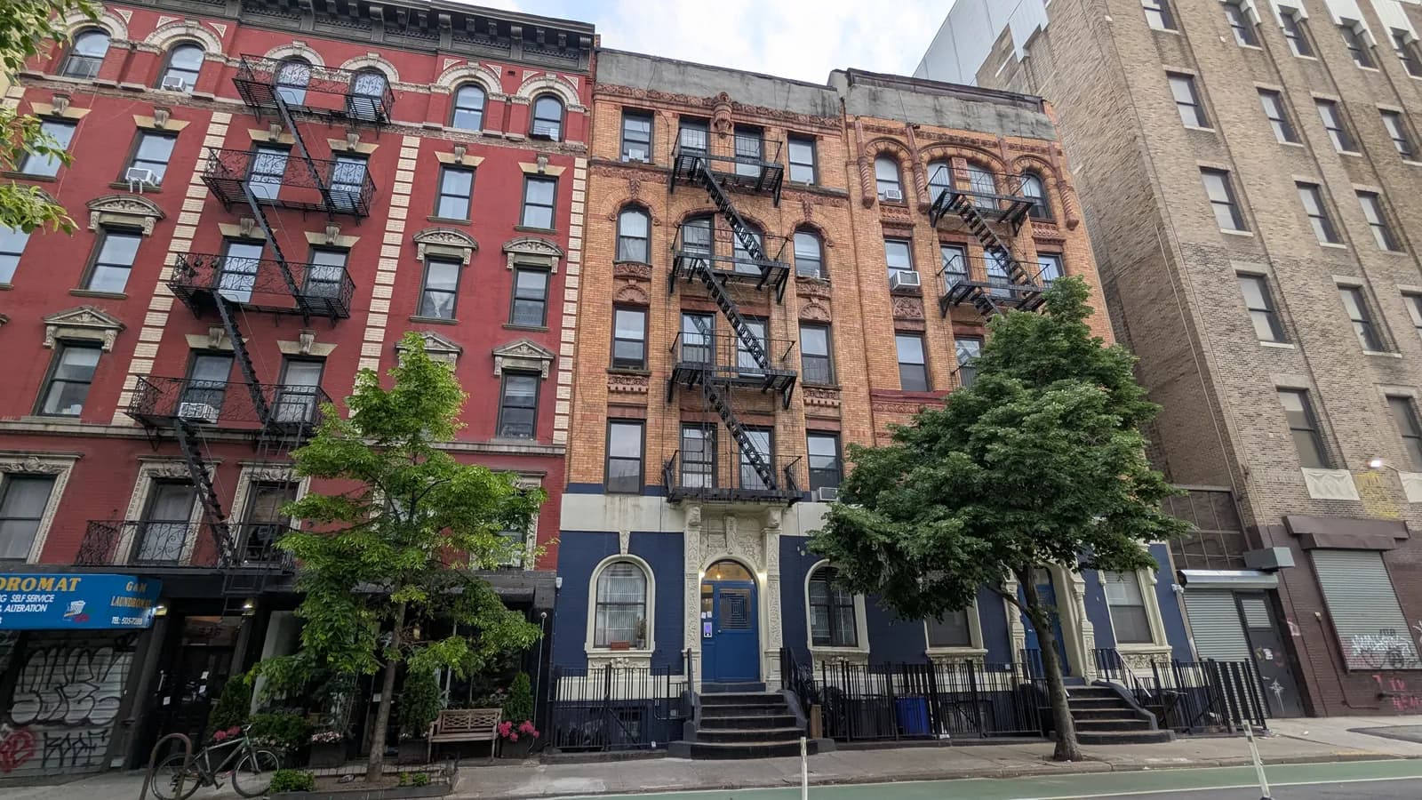 Five-storey walk-up apartment buildings in red-painted and orange brick on 13th Street in New York City, with black zigzag fire escapes, a stoop up to a blue front door, street trees and a laundromat at street level, beside a taller tan brick building.