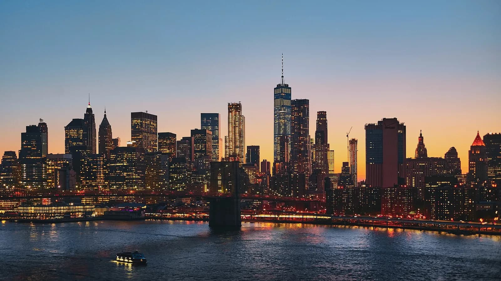 Lower Manhattan skyline at dusk seen across the East River, with One World Trade Center lit against an orange and blue sky, a span of the Brooklyn Bridge crossing in front, and a small lit boat on the dark water.