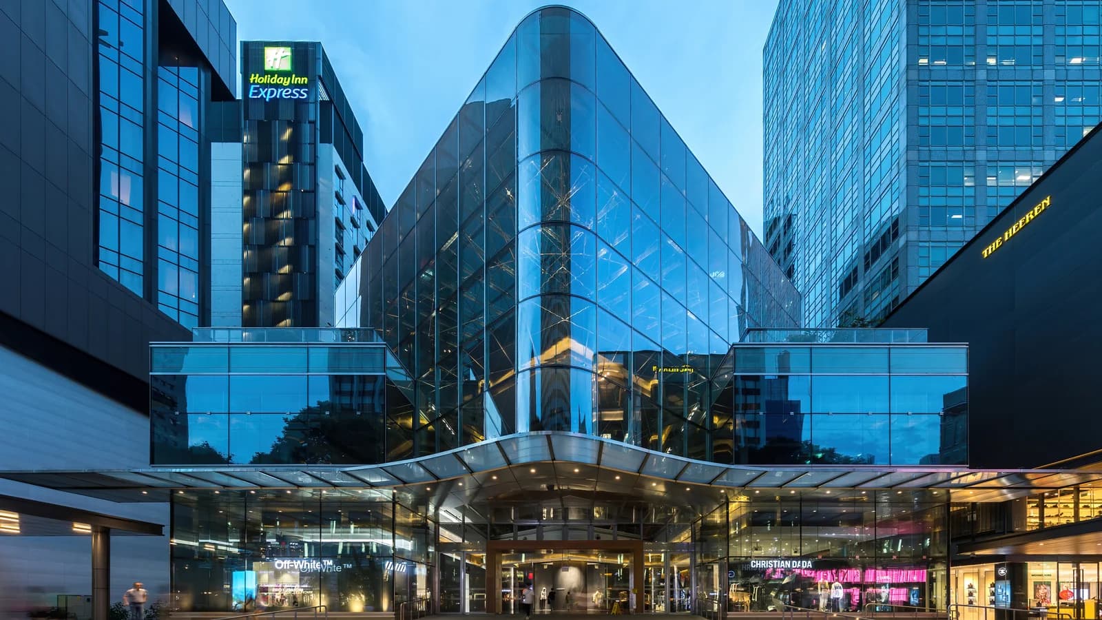 Illuminated glass facade of a shopping mall on Orchard Road at blue hour, with vertical reflections in the curtain wall.