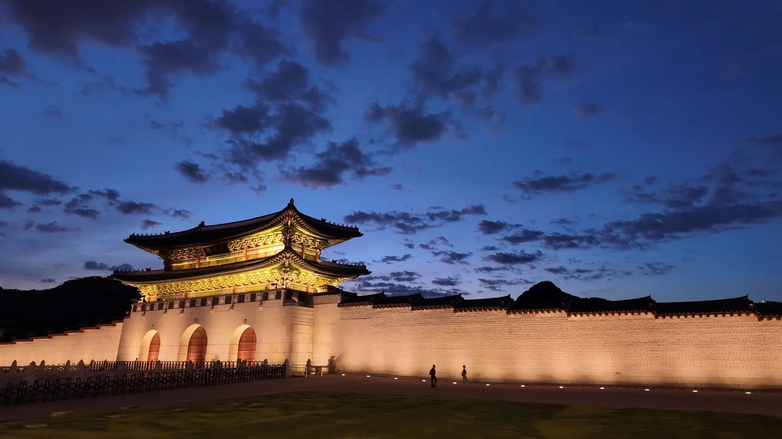 Gyeongbokgung Palace in Seoul lit at night, with traditional tiled roofs and wooden halls glowing under a deep blue evening sky.