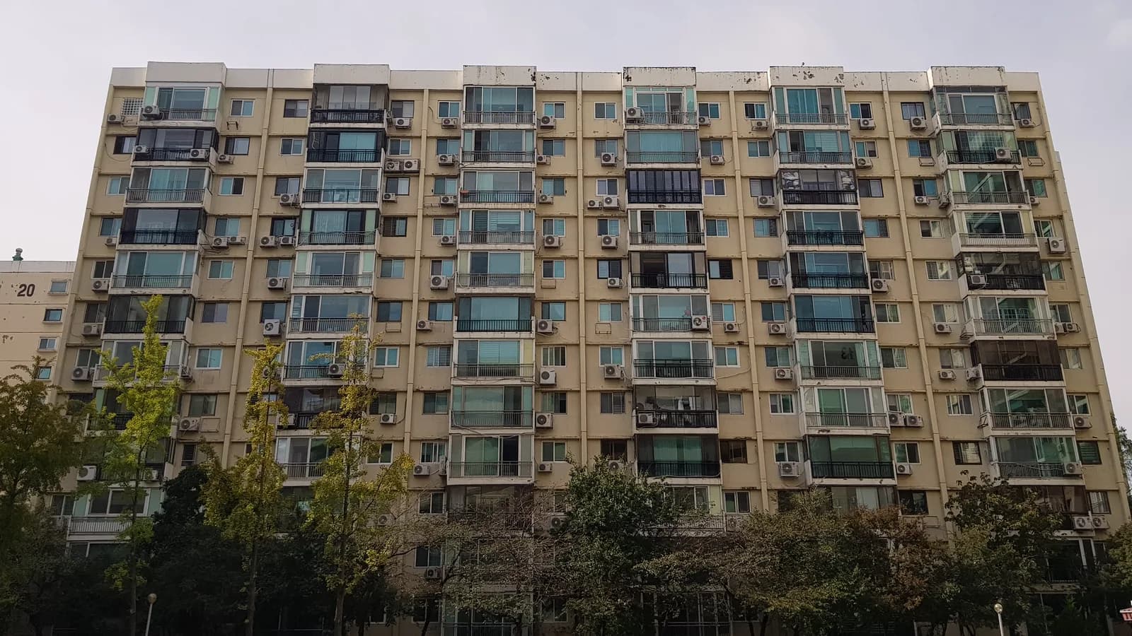 A long cream-coloured 1970s apartment block with rows of enclosed balconies and air-conditioning units, rising behind a line of trees under a pale sky in Apgujeong-dong, Seoul.