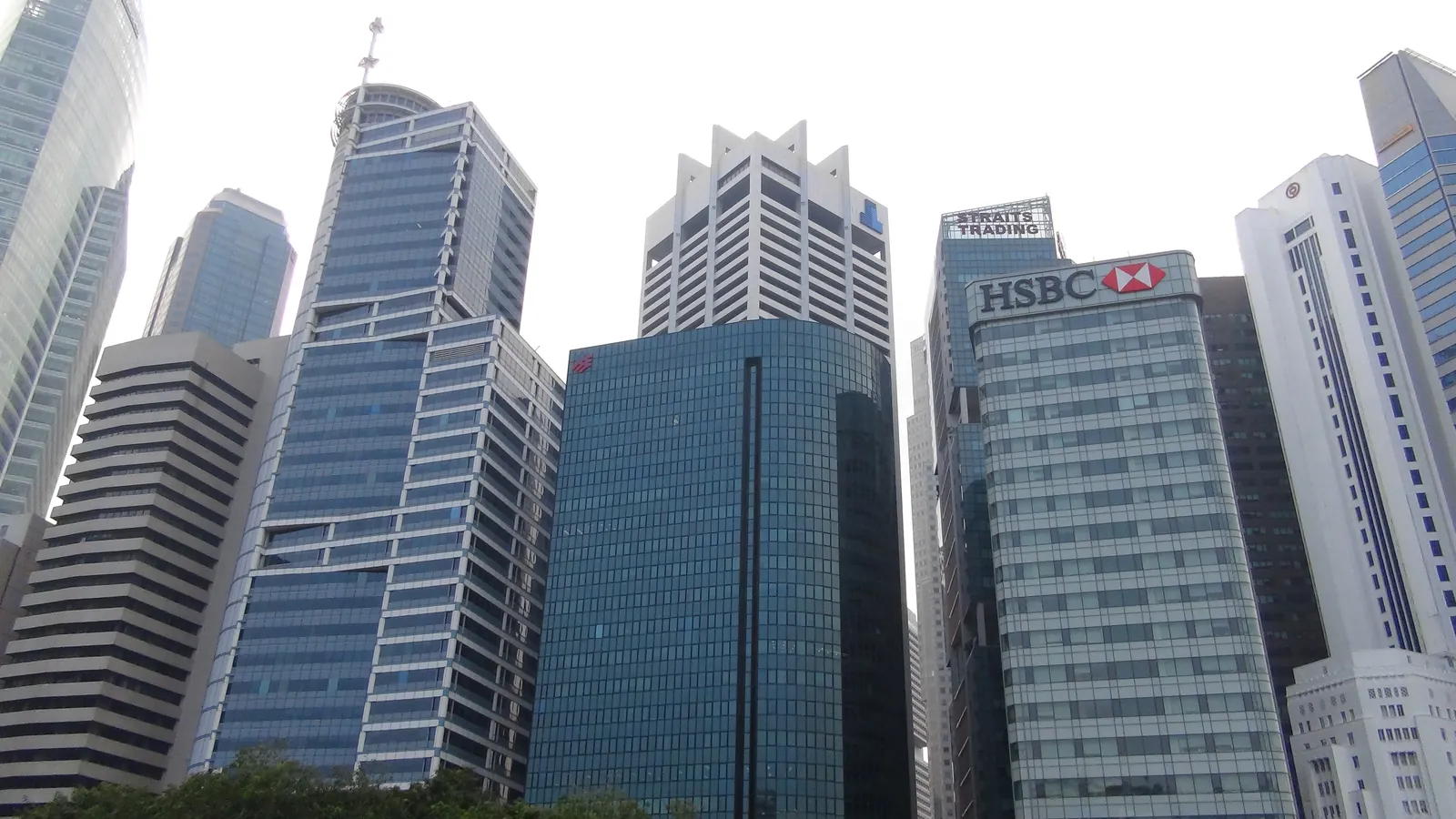 Office towers at Raffles Place in Singapore's Central Business District under a pale sky, including a blue-glass tower topped by a mast, a curved dark-glass tower and a building with an HSBC sign.