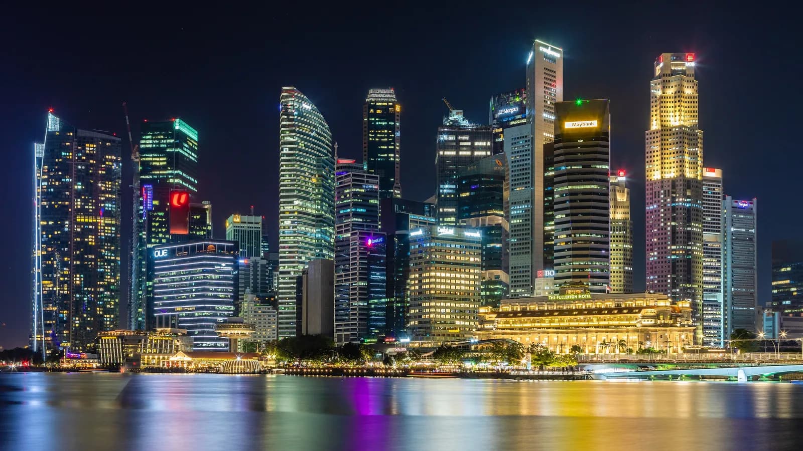 The Singapore Central Business District skyline at night, with lit office towers and the Fullerton Hotel reflected in the waters of Marina Bay.
