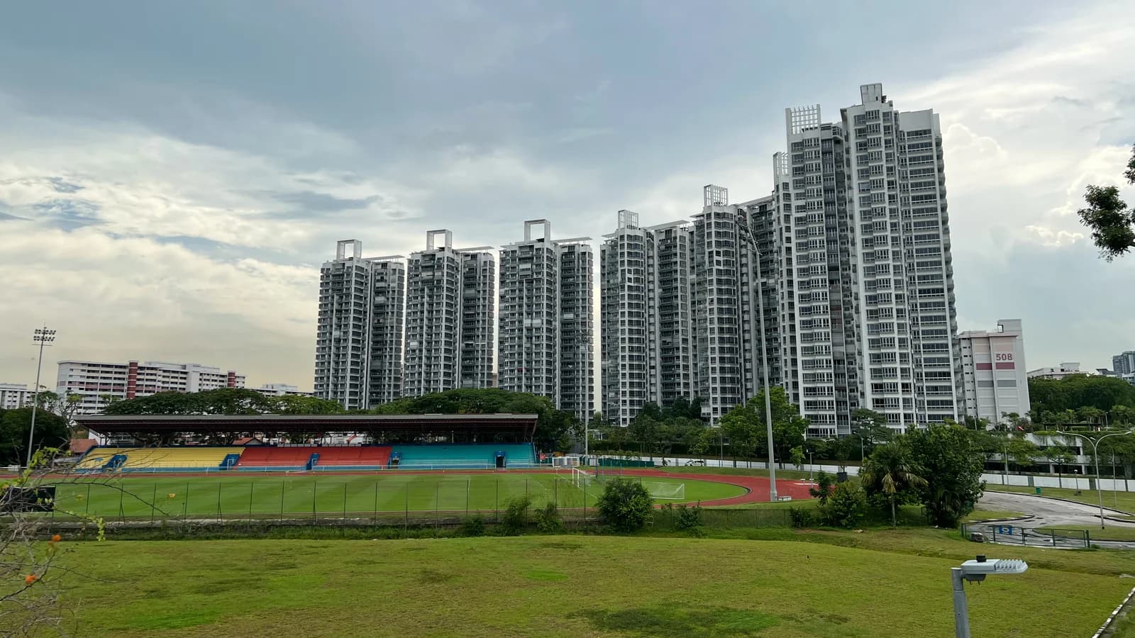 A cluster of white high-rise condominium towers under a cloudy sky, rising behind a stadium with a running track, a covered grandstand and a grass field, with lower housing blocks at the edges.