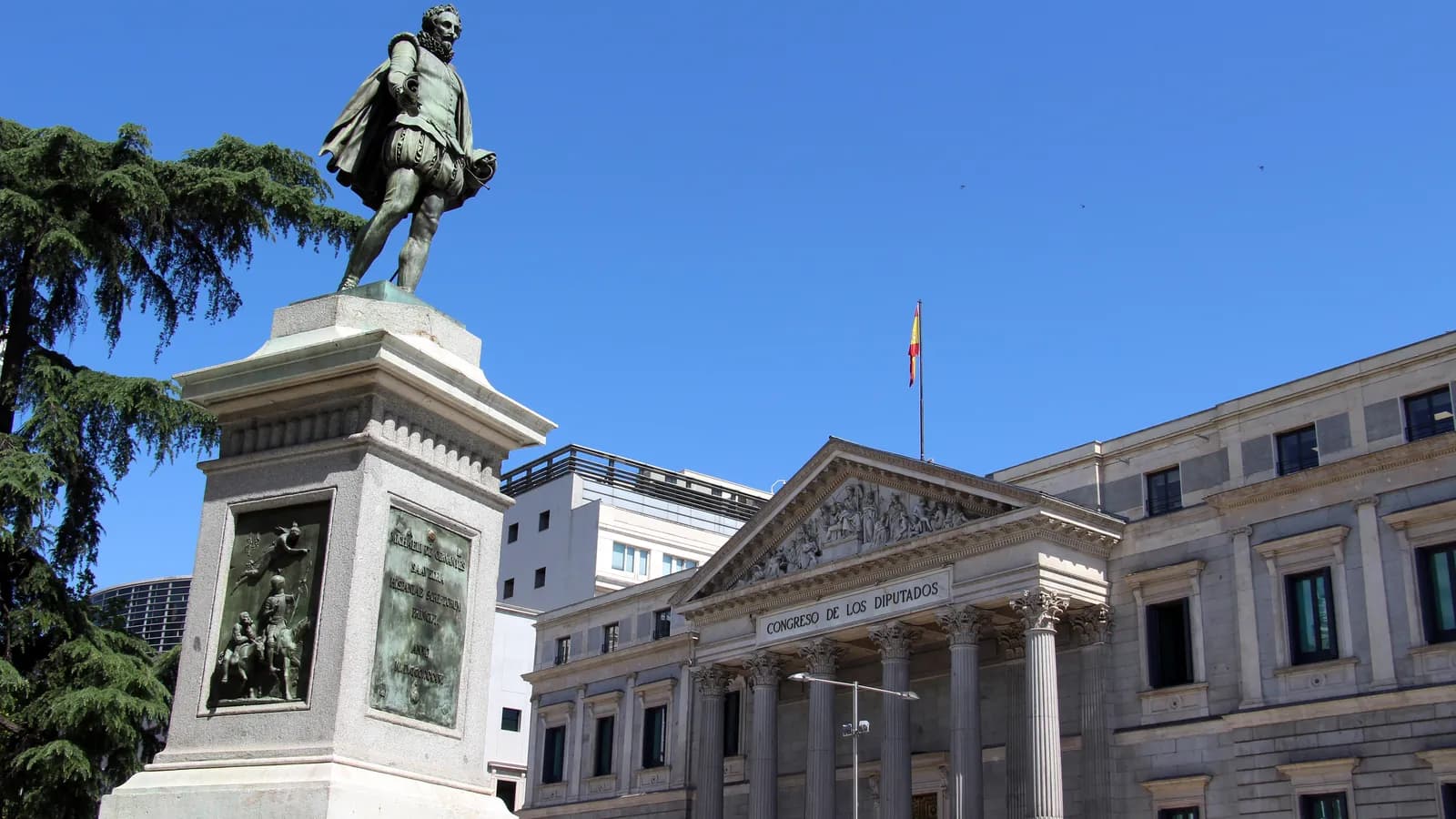 The columned front of Spain's Congress of Deputies building in Madrid, with the words Congreso de los Diputados above the entrance and a Spanish flag on the roof, behind a bronze statue of Miguel de Cervantes on a stone pedestal, under a clear blue sky.