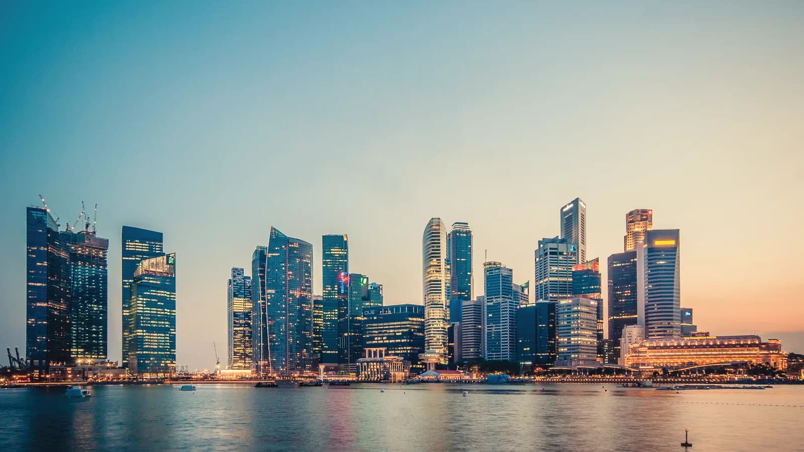 Singapore's Central Business District towers at dusk across Marina Bay, with office lights on and calm water in the foreground.
