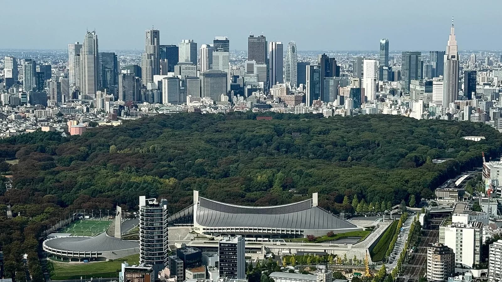 Shinjuku's high-rise skyline rising behind the forest of Yoyogi Park, with apartment and office blocks of Shibuya in the foreground, seen from above in Tokyo.
