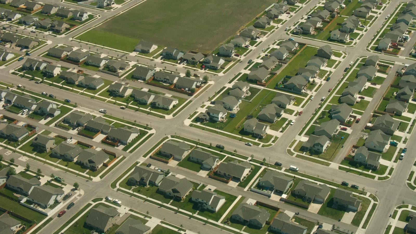 An aerial view of a newly built suburban development, with rows of grey-roofed houses along a grid of streets and a green field at the top.