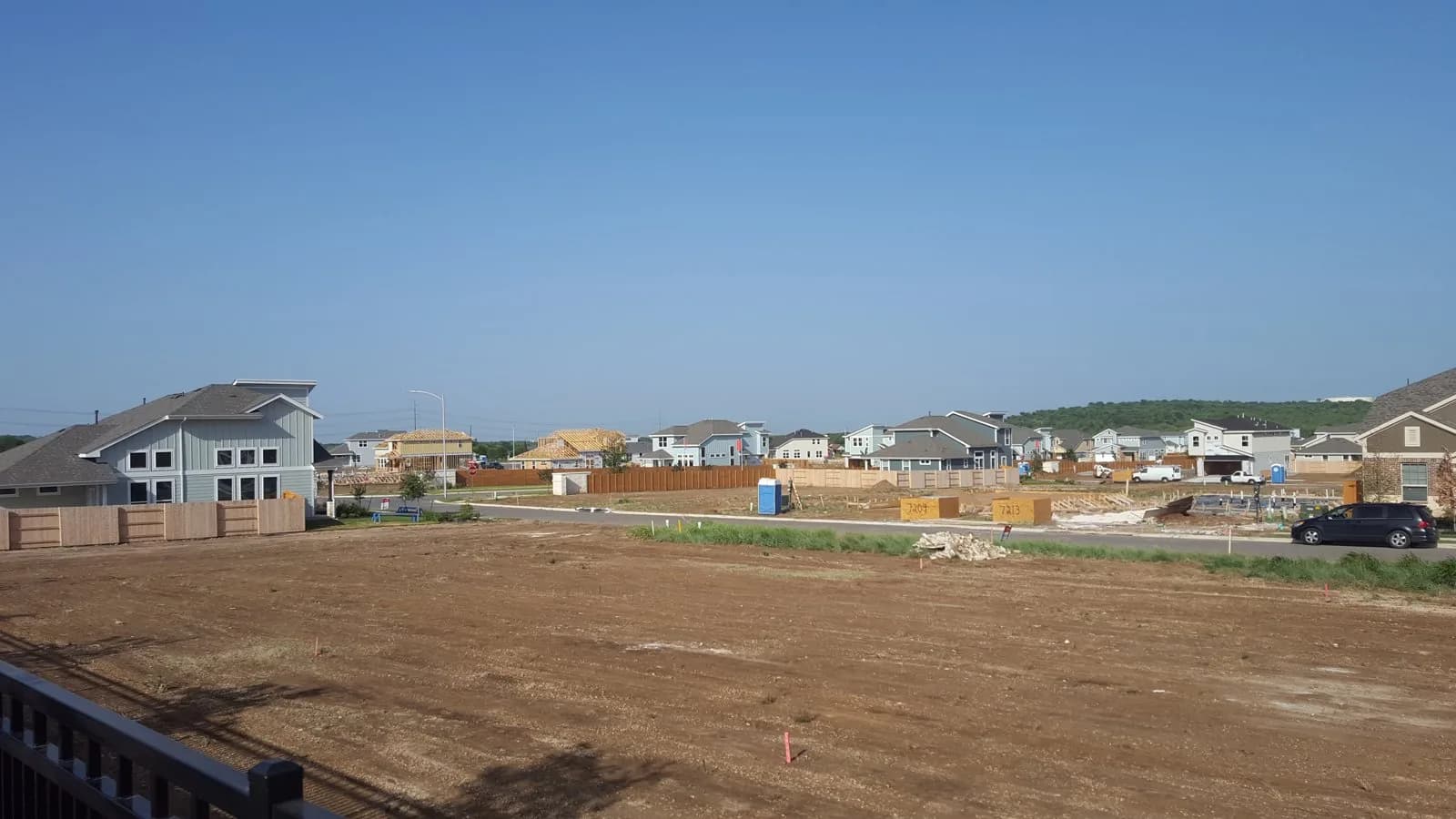 Cleared building lots in the foreground of the Easton Park development in Austin, Texas, with newly built two-storey houses and timber-framed homes under construction behind a fence line, under a clear blue sky.